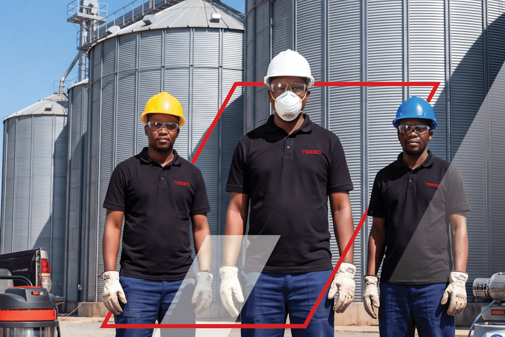 Three workers in hard hats and gloves stand in front of large silos at an industrial facility.